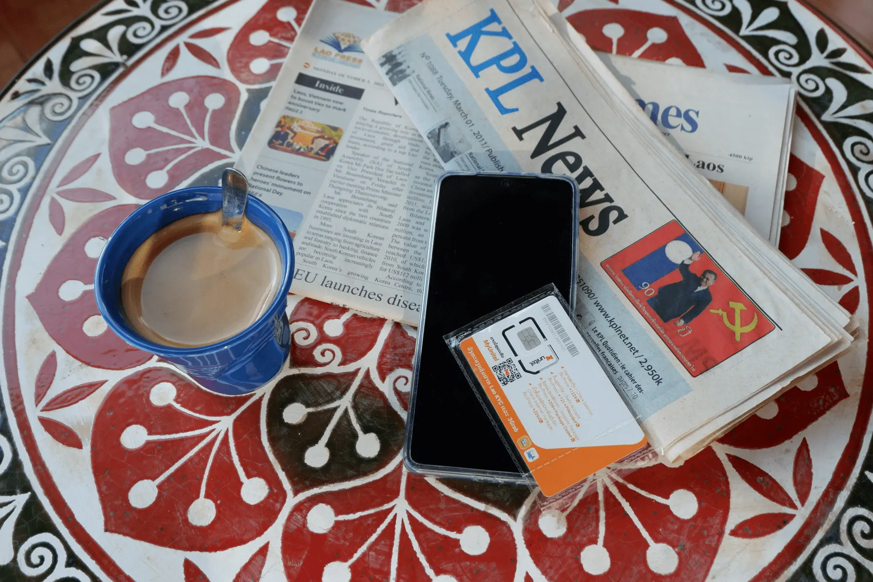 Illustration générée par IA : tasse de café, journaux KPL, un téléphone et une carte SIM sur une table à motifs rouges